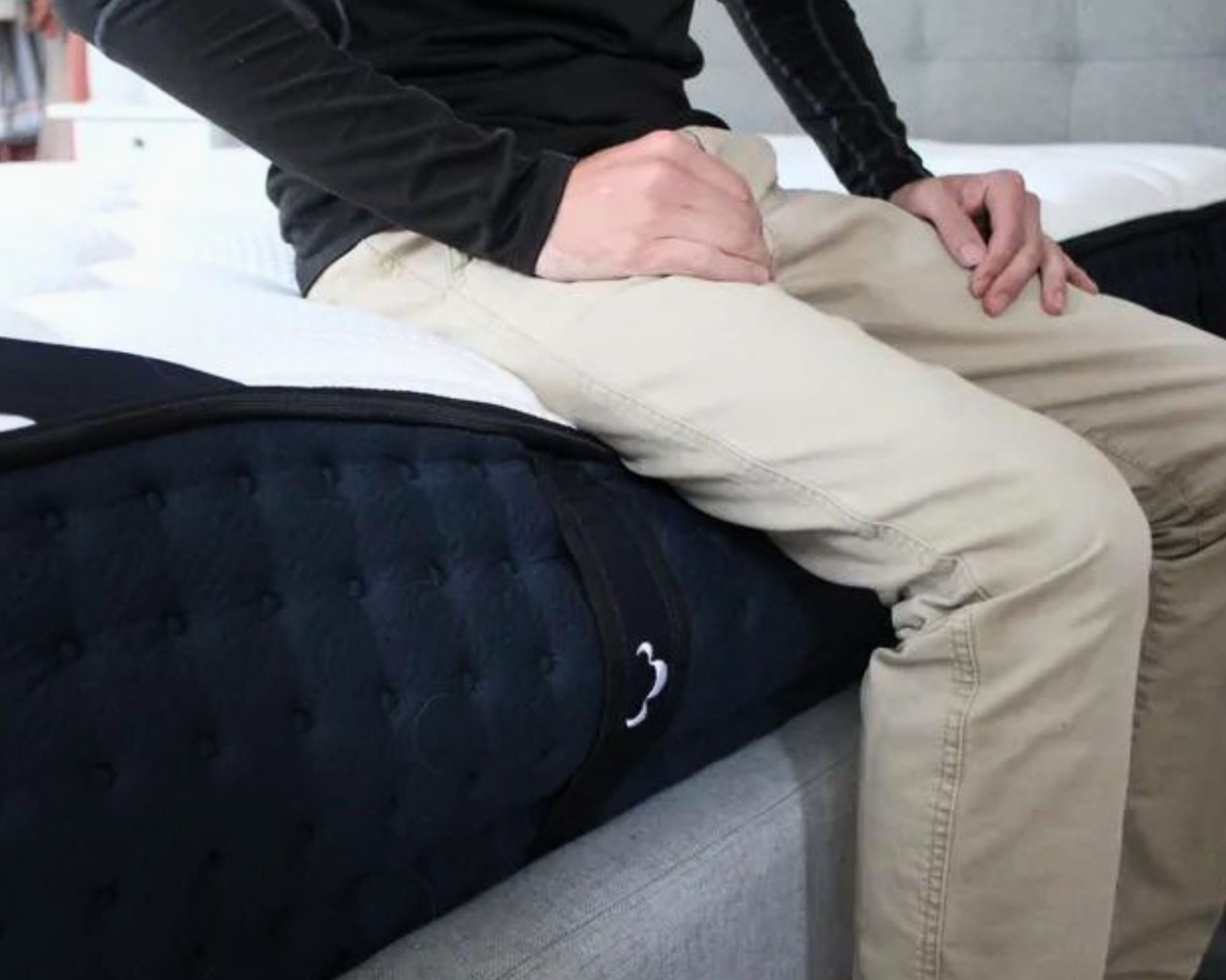 A close-up of a man wearing a black long-sleeved top and beige trousers sitting on the end of the DreamCloud Classic Hybrid Mattress on a grey upholstered bedframe.