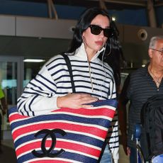 Dua Lipa walks through the airport carrying an oversized Chanel tote bag wearing a striped knit with jeans and black pointed-toe heels.