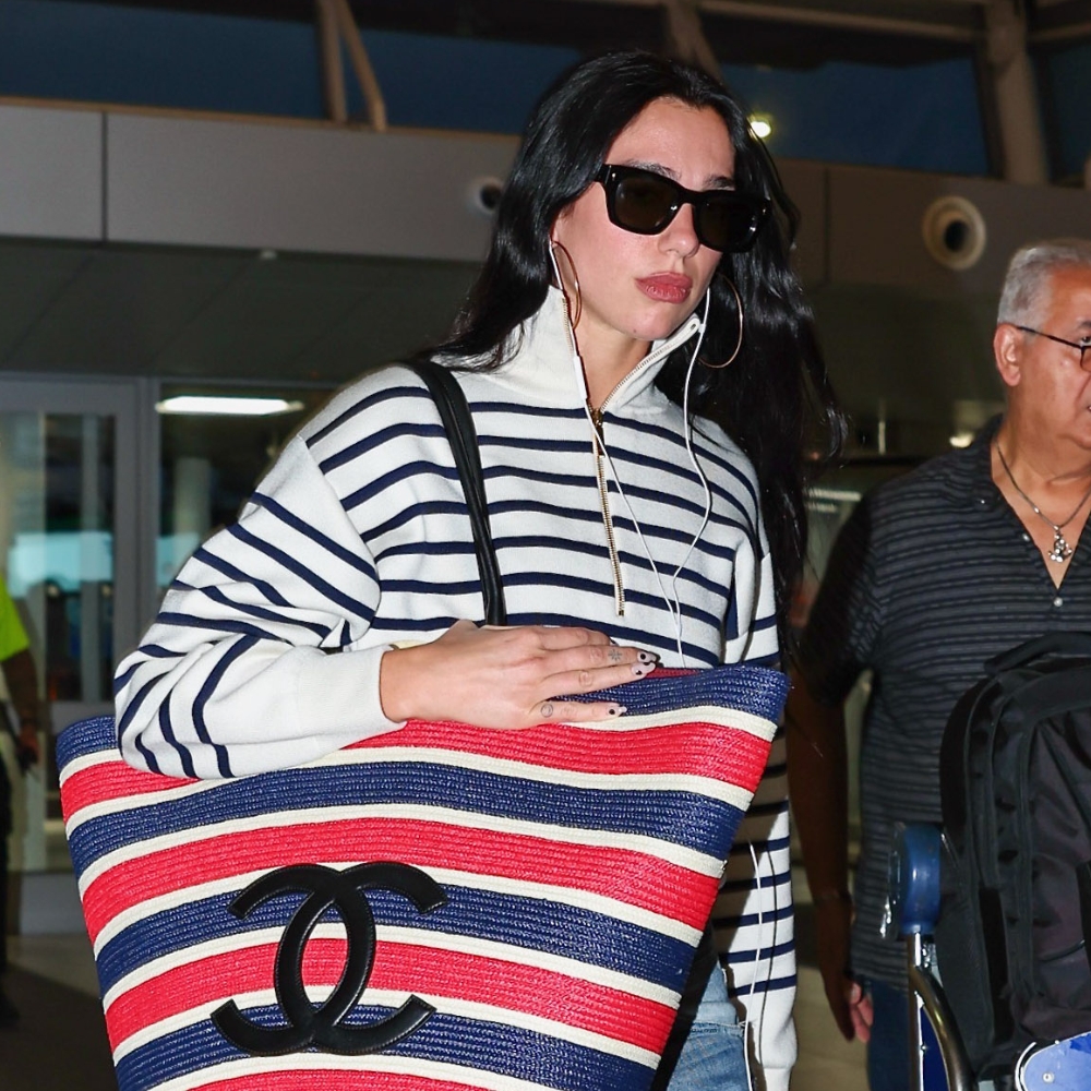 Dua Lipa walks through the airport carrying an oversized Chanel tote bag wearing a striped knit with jeans and black pointed-toe heels.