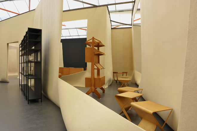 Inside an exhibition area featuring wood stools, a set of shelves in black and a unique shelf storage structure in brown/wood.