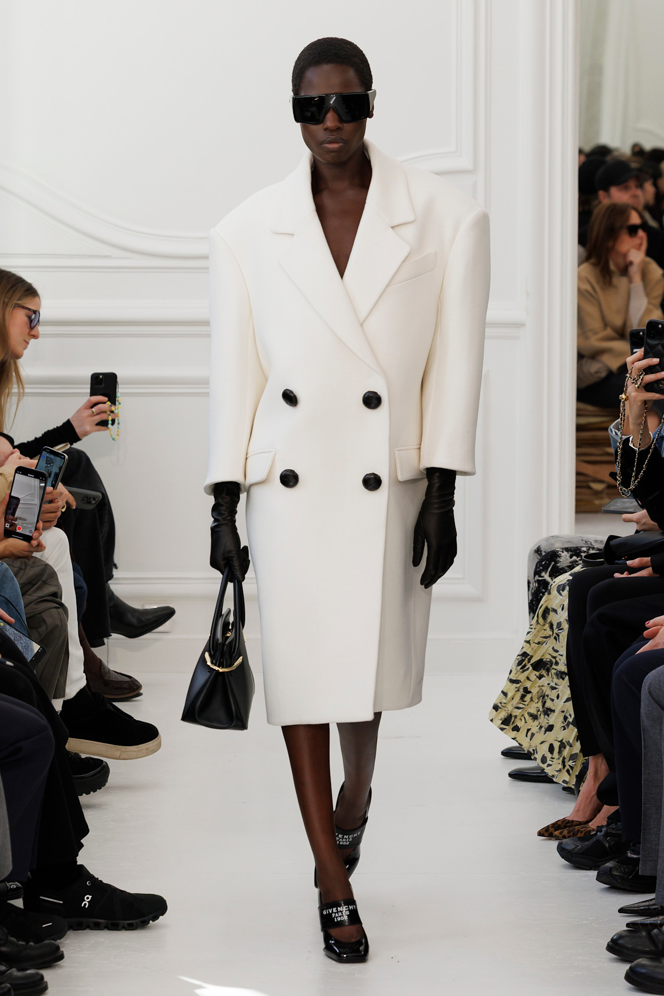A model wears a large white coat with rounded shoulders and black buttons on Givenchy&#039;s autumn/winter 2025 runway
