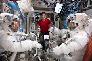 two astronauts in spacesuits on either side of picture with an astronaut floating without a spacesuit in between. the astronaut on the right with a spacesuit gives a thumbs-up