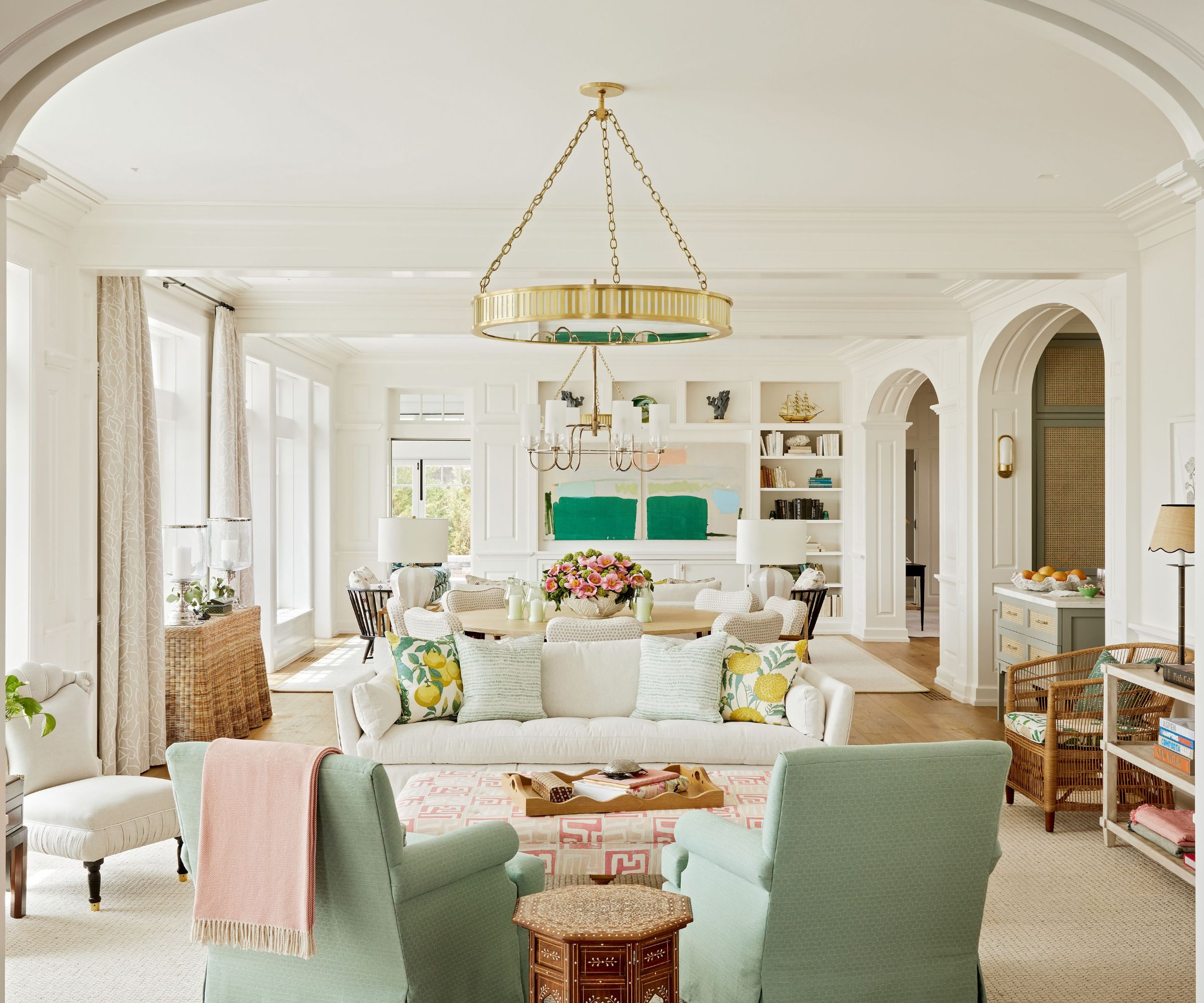 Grand living room with white walls, high ceilings, arched doorways, a large sofa in the middle with an upholstered ottoman, two pale green armchairs, built-in bookshelves and a dining table