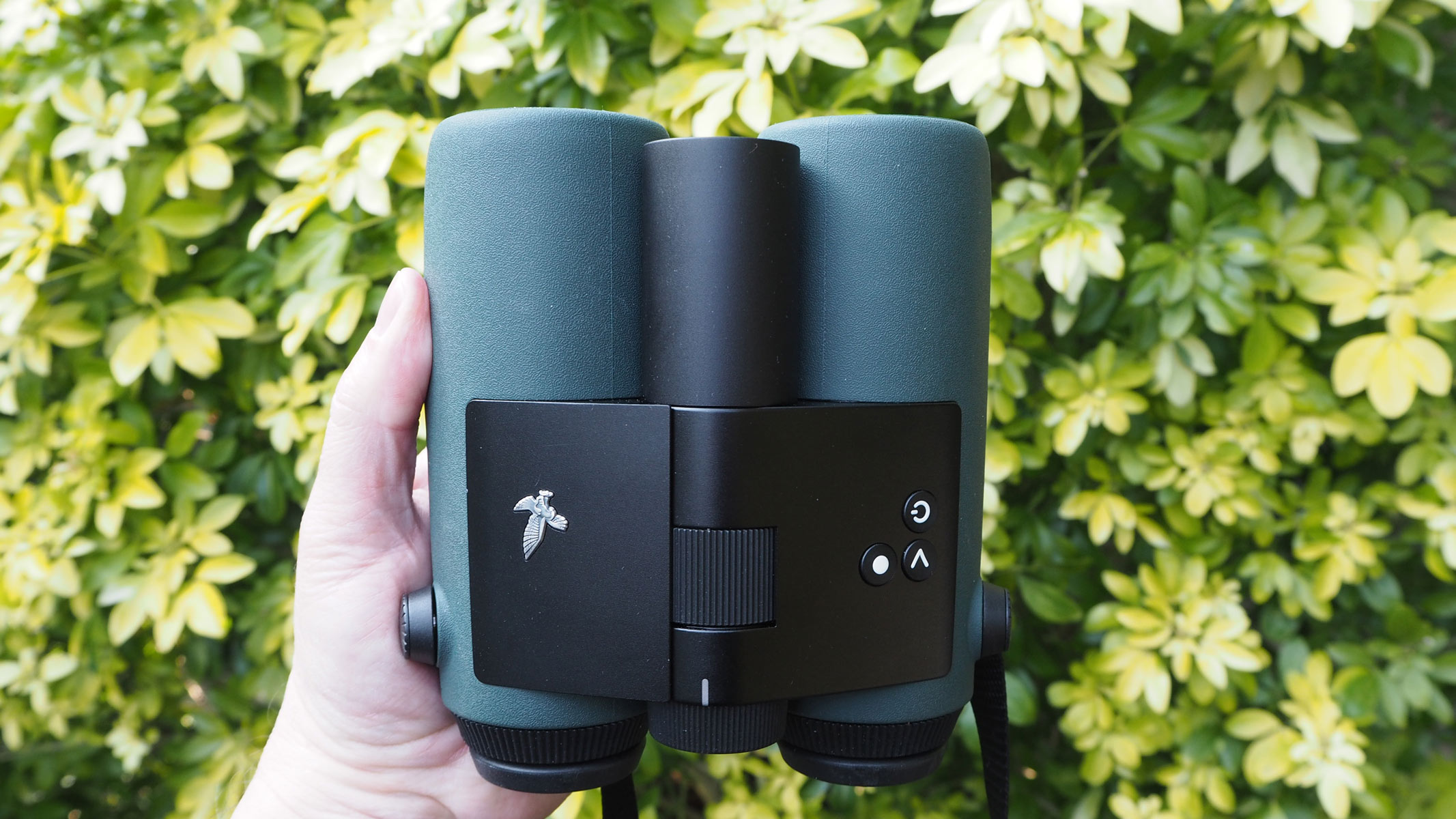 A hand holding the Swarovski AX Visio binoculars against a background of bush leaves.