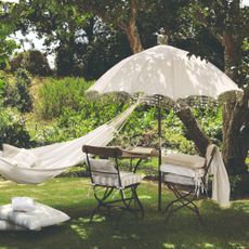 An English garden with a fringe-trimmed white parasol, a bistro set underneath it and a white hammock tied to the tree next to it