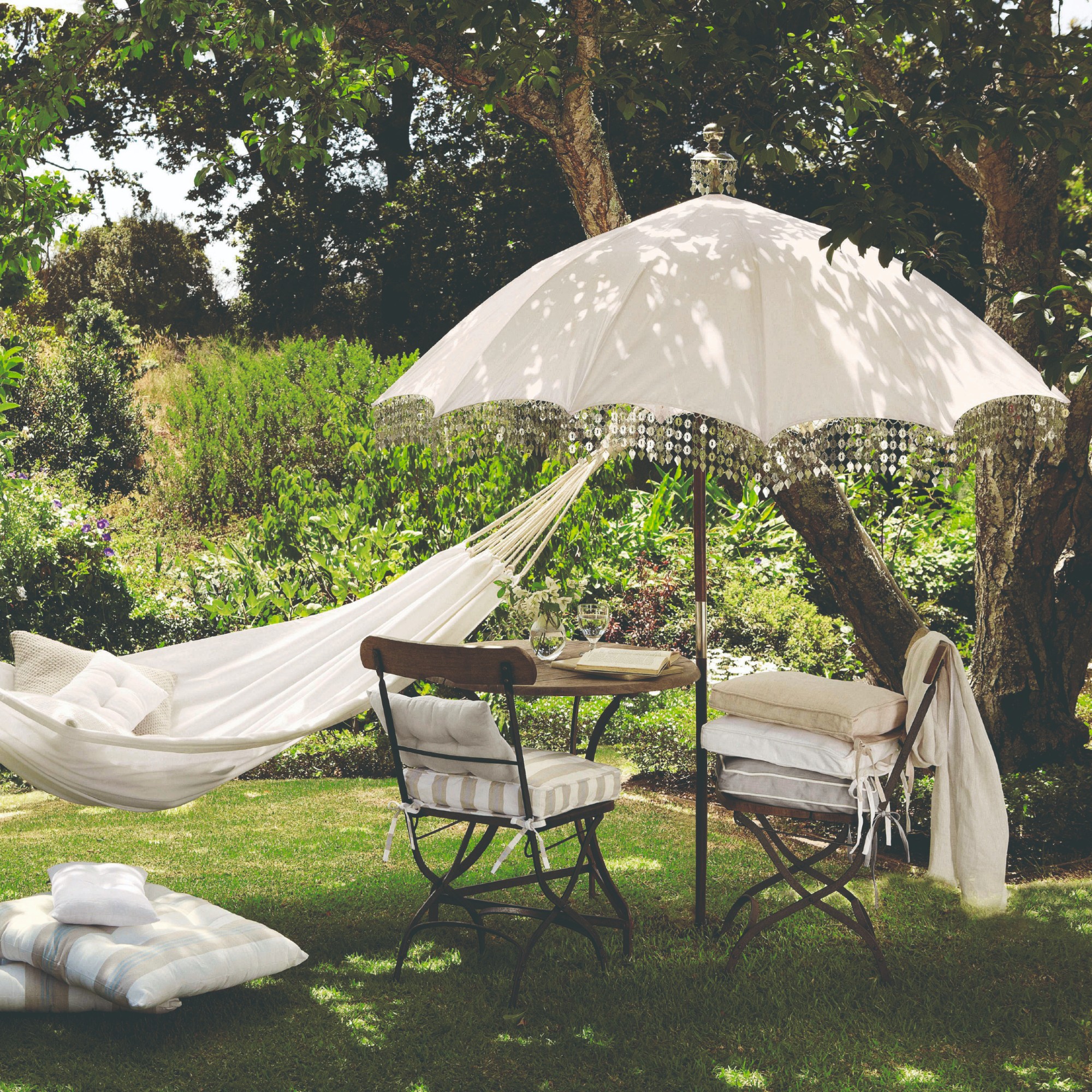 An English garden with a fringe-trimmed white parasol, a bistro set underneath it and a white hammock tied to the tree next to it