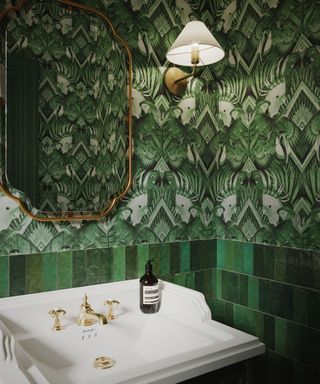 A dark green bathroom with dark green tiles and floral wallpaper, a gold mirror, a white wall sconces, and a white basin with a gold faucet