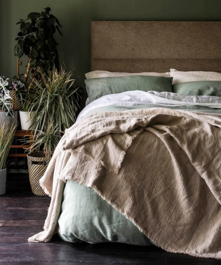 A light beige bed frame with light green bedding and a tetxured beige throw next to multiple houseplants