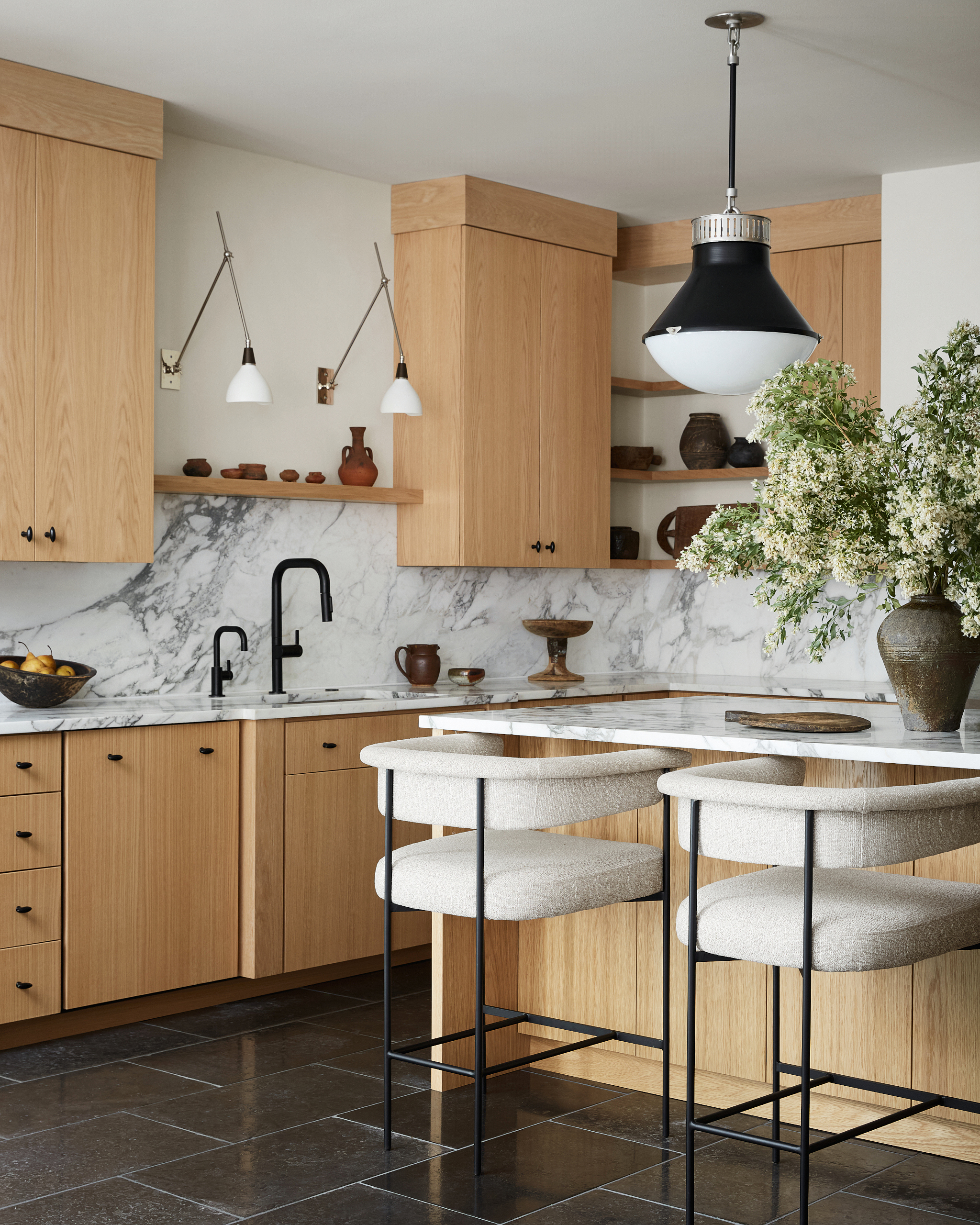 Oak kitchen cabinetry and white marble splashback with black tiled floor, white upholstered bar stools with thin black metal frames, and black pendant light