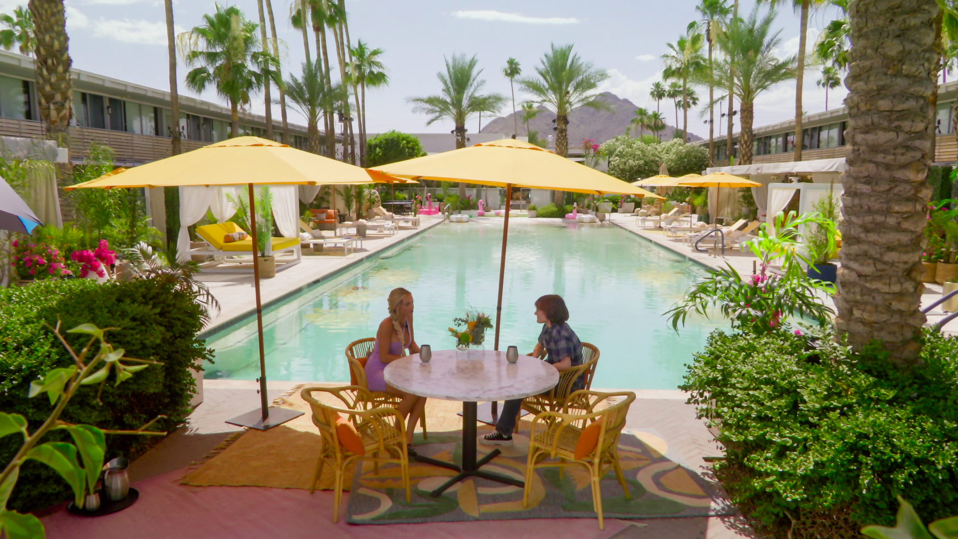 Two people sit at an outdoor table in front of the pool at a motel, with multiple palm trees and a desert mountain in the distance, in 'Sneaky Links.'