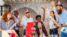 Big group of people celebrating a birthday. There are 6 people in total, all cheering and throwing confetti in the air. Three are wearing party hats. In front of them is a table with birthday decorations, such as a chocolate cake and drink cups.