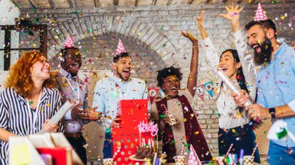 Big group of people celebrating a birthday. There are 6 people in total, all cheering and throwing confetti in the air. Three are wearing party hats. In front of them is a table with birthday decorations, such as a chocolate cake and drink cups.