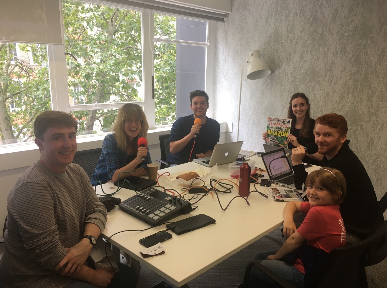 A mixed group of adults and children sit around a table with microphones and computers recording a podcast.