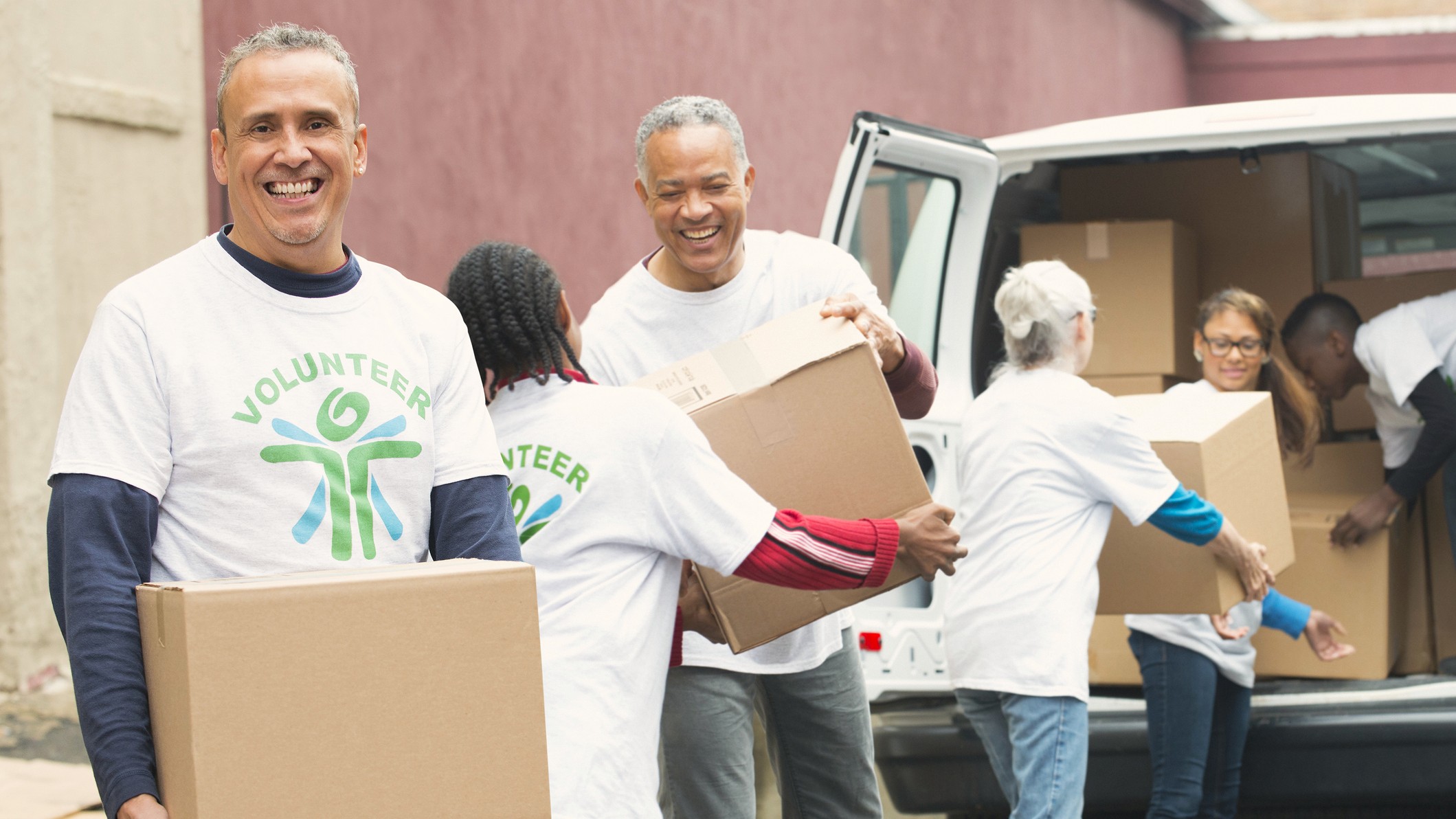 An older volunteer smiles at the camera. He and others are moving boxes out of a truck. They wear tee-shirts identifying themselves as volunteers.