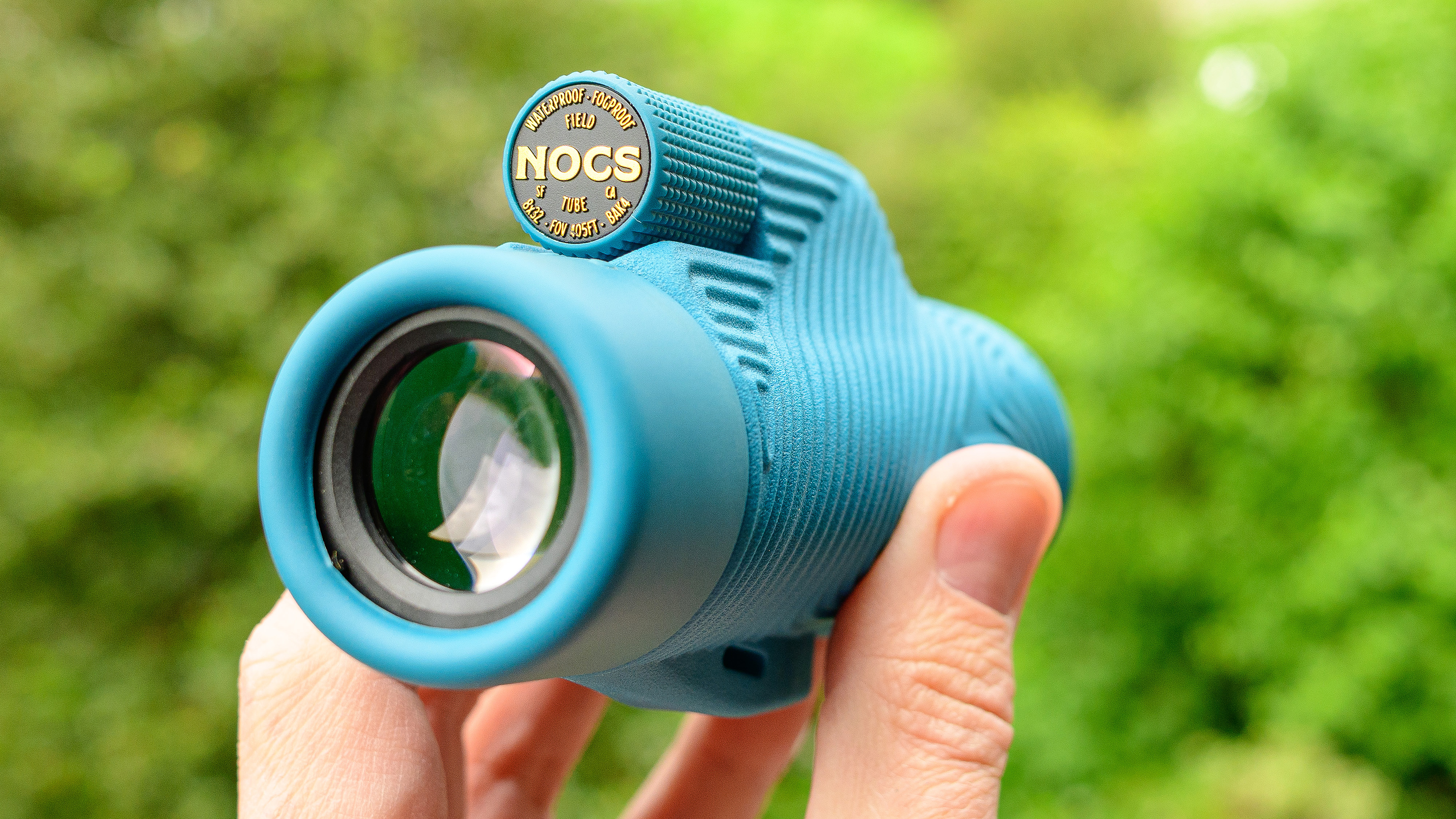 Nocs Field Tube monocular in blue.  Depicted on the user's hand against a green background.