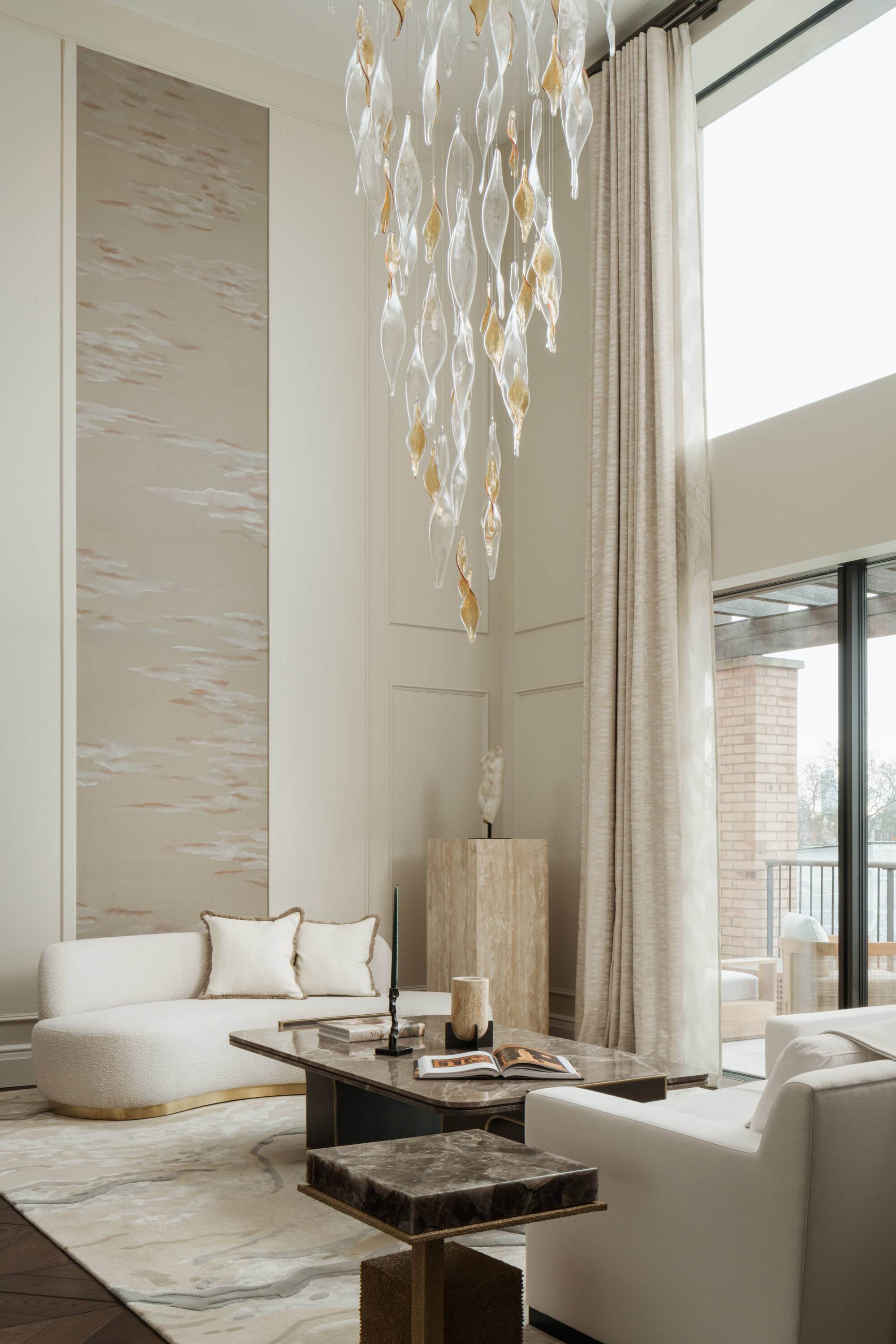 beige living room with glamorous furnishings, wall moldings, white sofa, glass chandelier, white patterned rug, marble coffee table, travertine plinth, and double-height ceilings