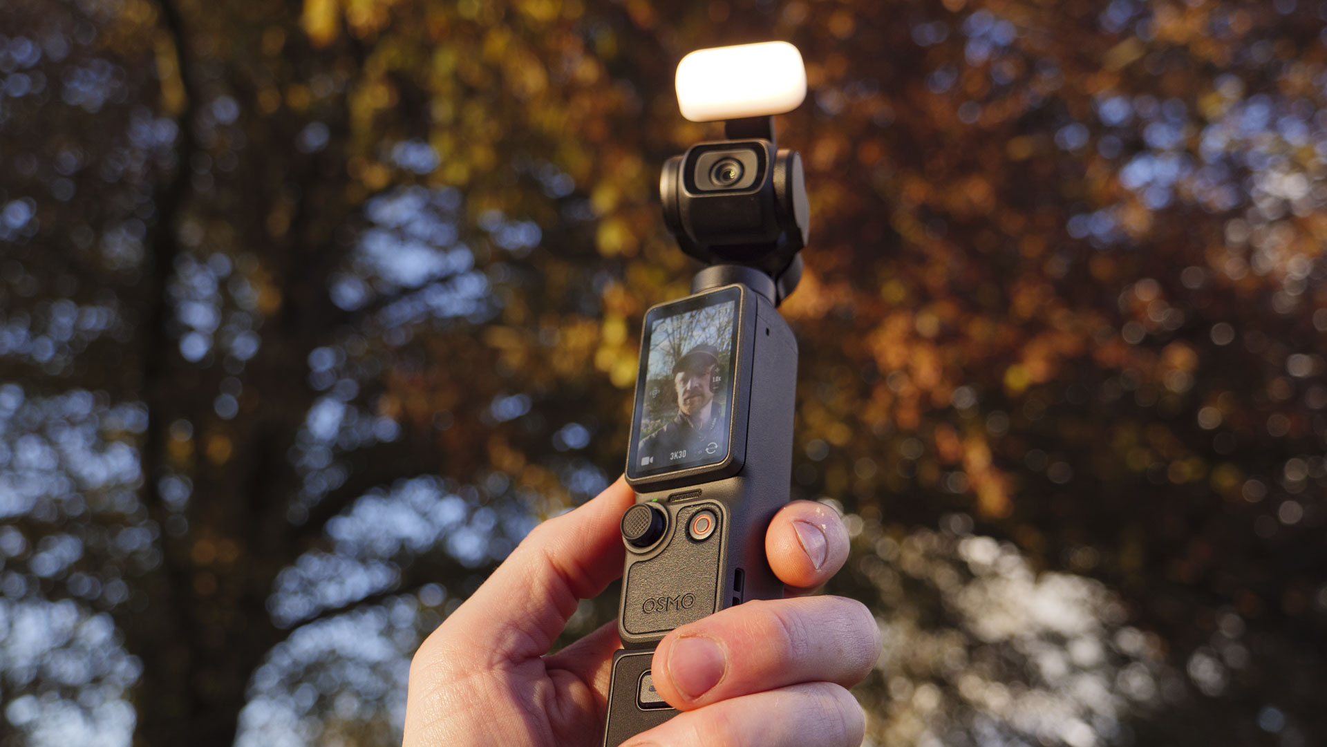 DJI Osmo Pocket 4 vlogging camera with natural background at golden hour, and the user in the frame of the screen