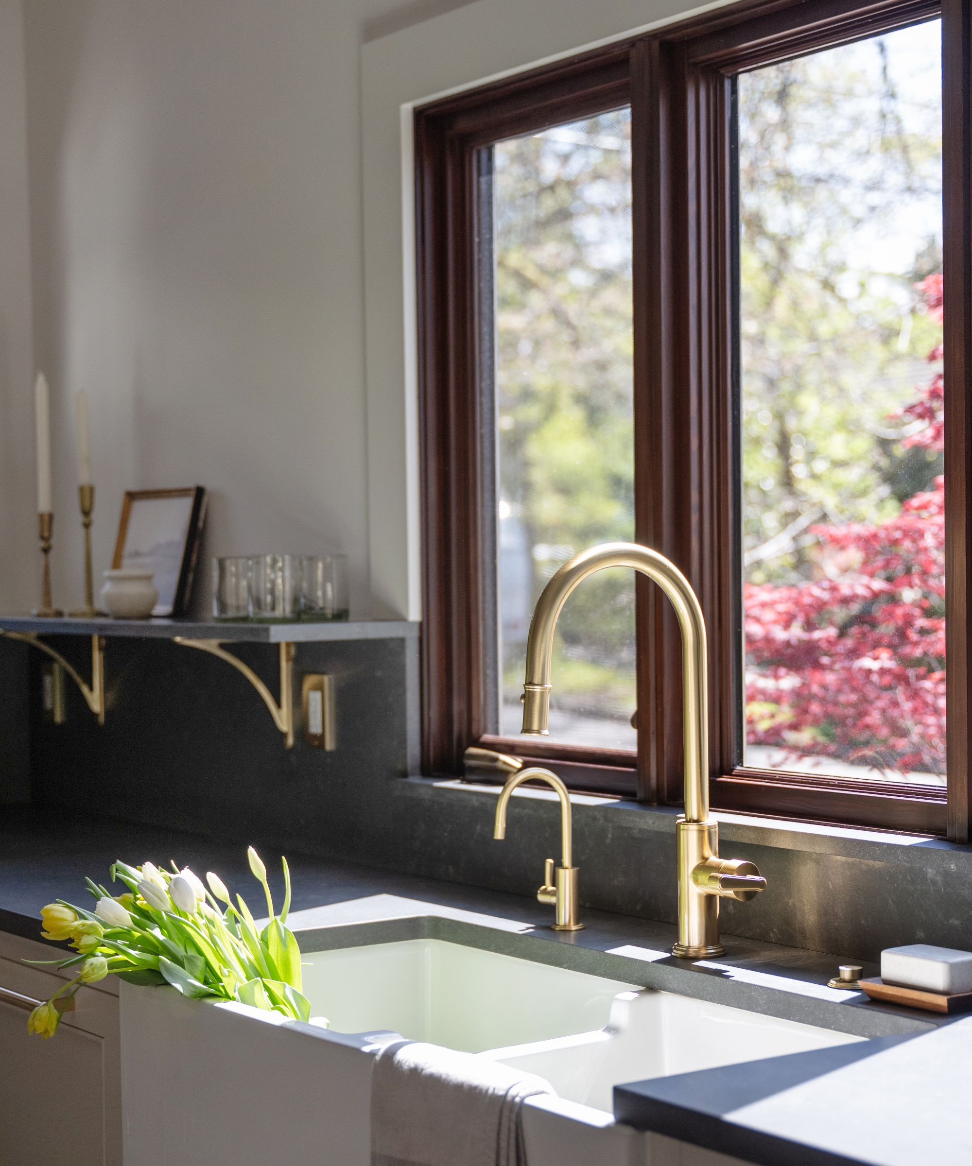 A close-up of a kitchen sink installed beneath the window, with a brass faucet and black countertops surrounding it