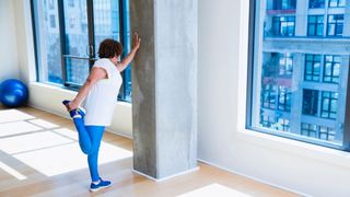 Woman holding onto a wall with one hand and stretching a leg with the other, representing wall Pilates