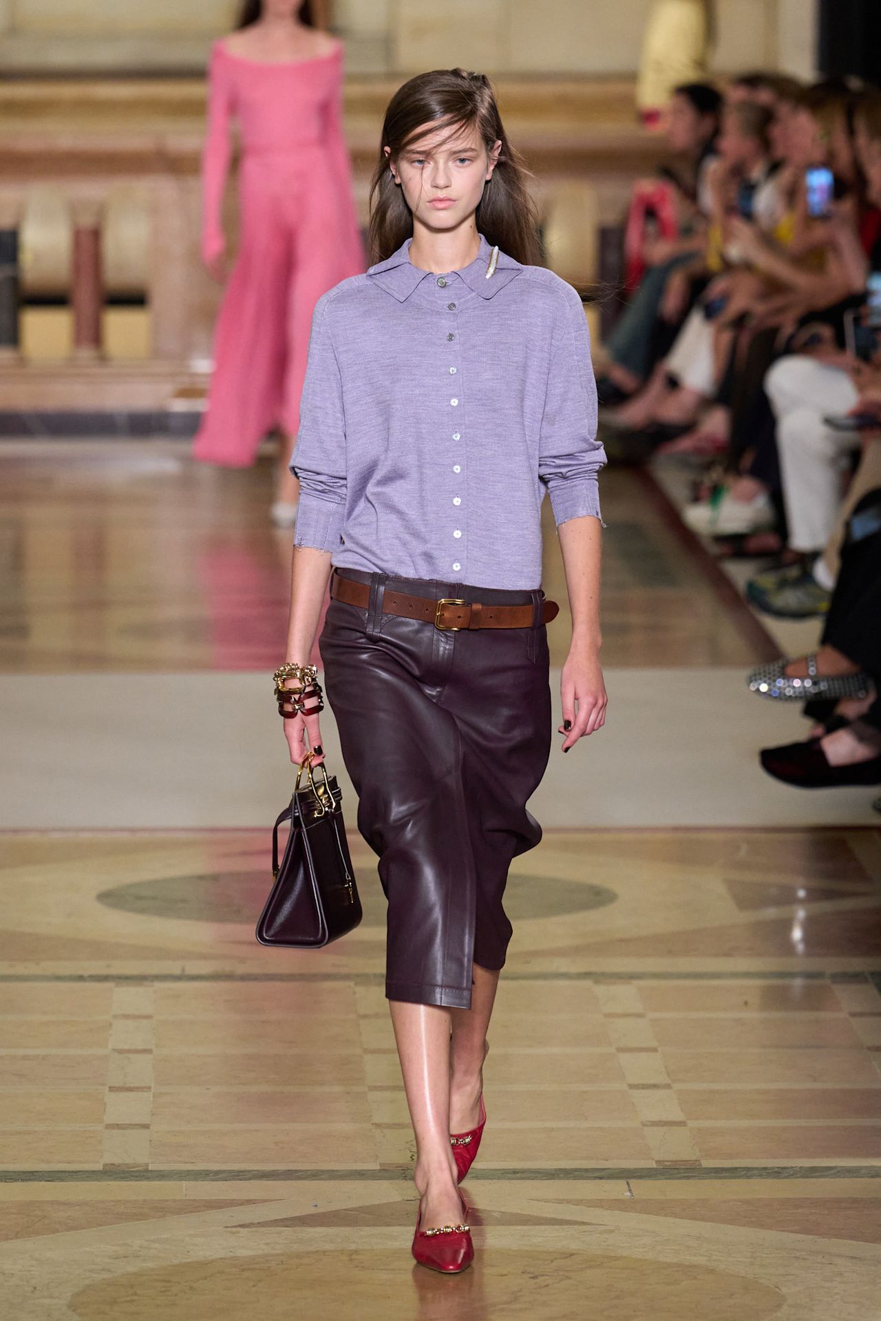 A Tory Burch model wearing a light purple polo layered underneath a matching cardigan, an aubergine-hued leather pencil skirt, a brown leather belt, stacked chain bracelets, a brown structured bag, and red pointed pumps.