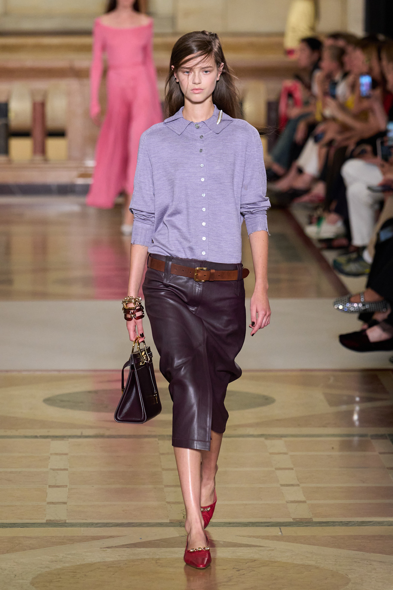 A Tory Burch model wearing a light purple polo layered underneath a matching cardigan, an aubergine-hued leather pencil skirt, a brown leather belt, stacked chain bracelets, a brown structured bag, and red pointed pumps. 