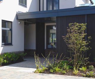 modern house with black porch extension, planted border around side of house and larger planted section in front of porch, edging onto paved driveway