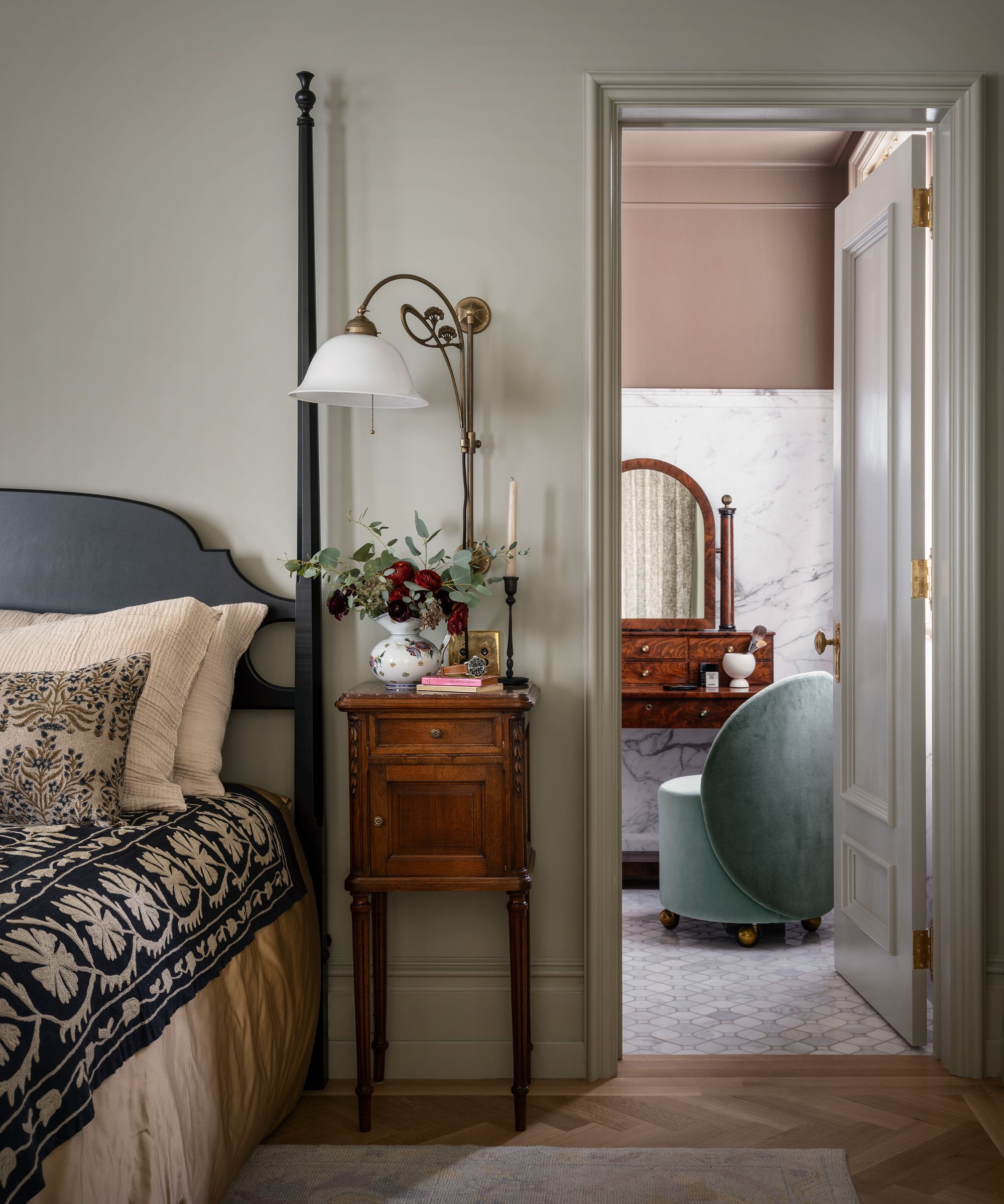 Bedroom with light gray-green walls, with a door leading into a bathroom. Parquet wood flooring, a black headboard with soft pink patterned bedding, a small wooden nightstand, and a brass wall sconce.