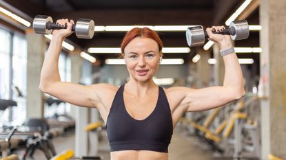 Attractive red-haired fitness woman trains shoulder muscles doing overhead dumbbell press in a modern gym. Bodybuilding and Fitness