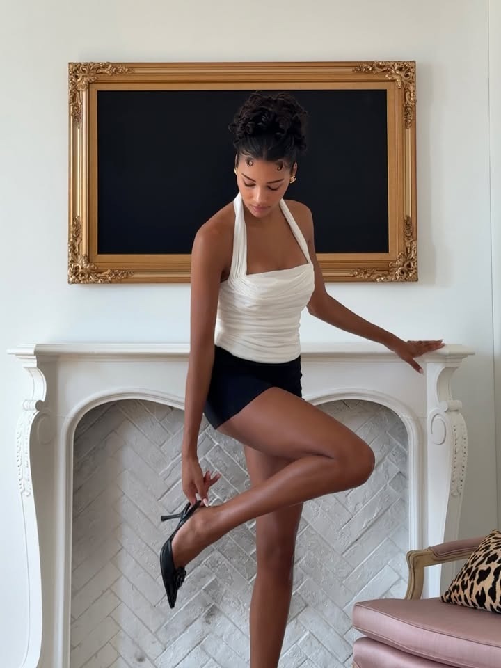 A woman leaning against a fireplace fixing her shoe.