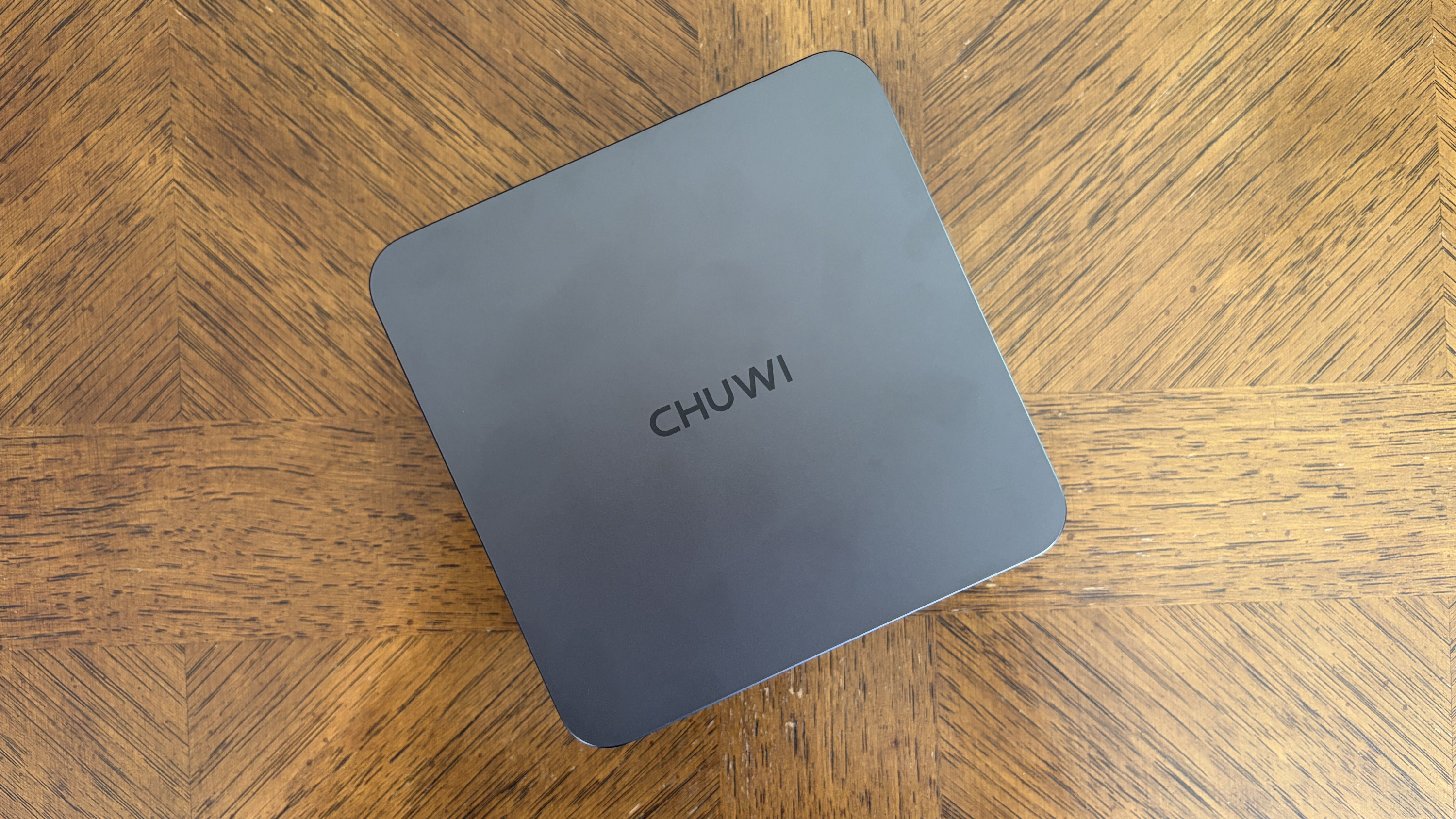 A square dark gray device with "CHUWI" on top, resting on a brown herringbone-patterned wooden surface.