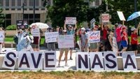 People hold signs outside on a sunny day in protest of NASA budget cuts, July 20, 2025.