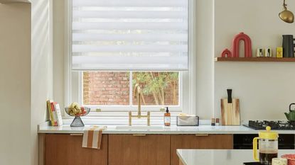 Image of a white, modern kitchen. There is a direct view of the kitchen sink, which has white countertops, gold hardware, and wooden cabinetry. There is a window above the sink with day and night blinds.