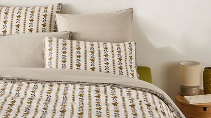 a bed made with reversible bedding with folksy print on one side and beige stripe on the reverse, timber side table with lamp, book, and vase