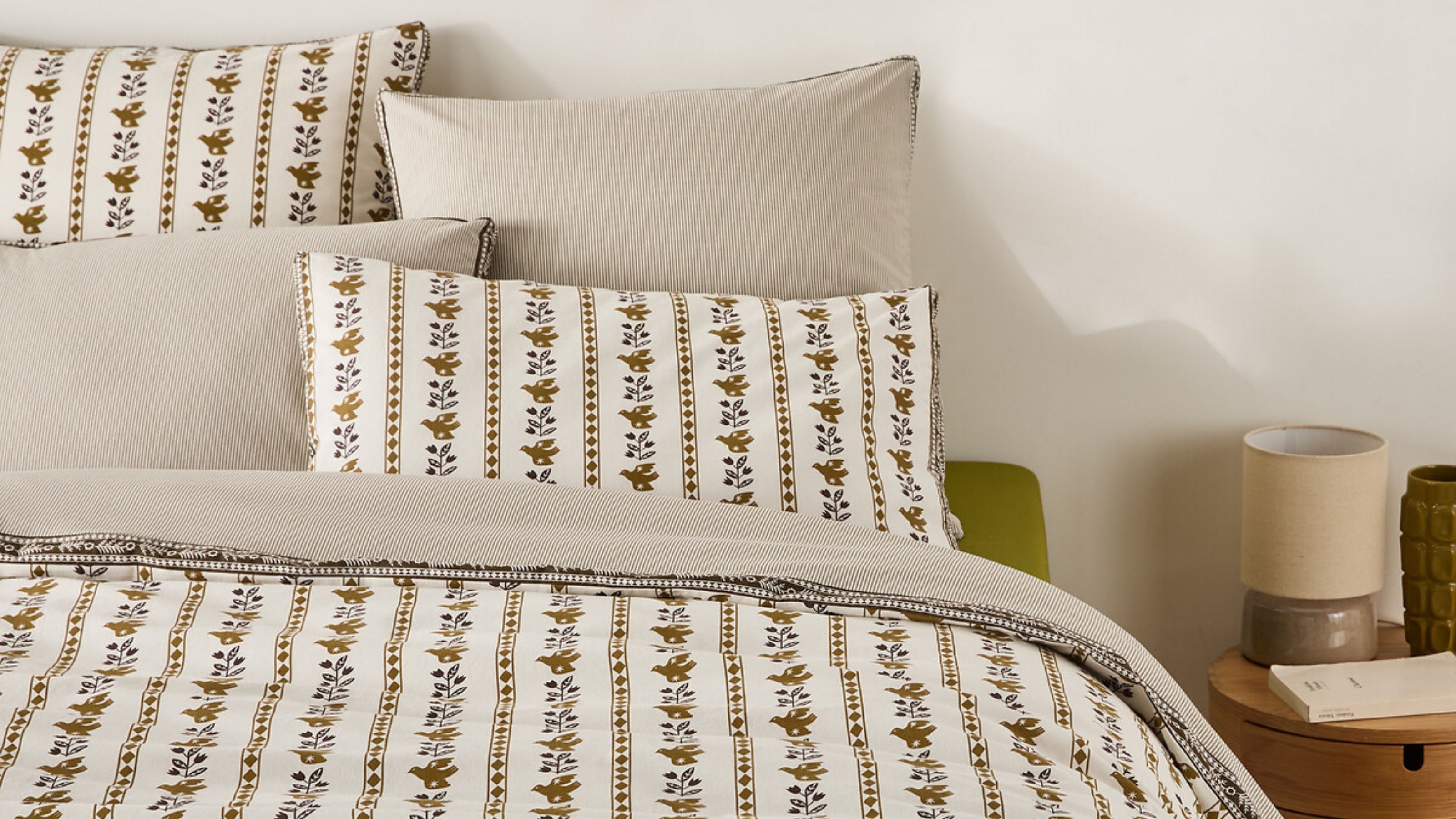 a bed made with reversible bedding with folksy print on one side and beige stripe on the reverse, timber side table with lamp, book, and vase