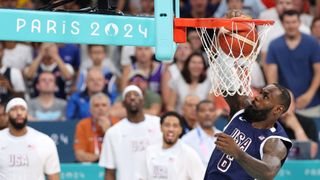 LeBron James for team USA Basketball is shown dunking the ball into the net during a game at the 2024 Paris Olympics while spectators and fellow team mates (who are blurred in the background) look on. 