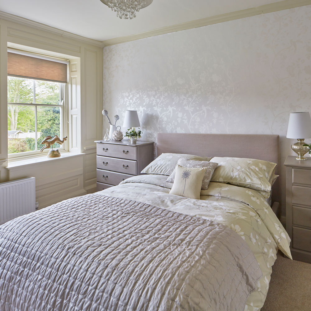 Bedroom with patterned wallpaper, double bed, side table with drawers and window.