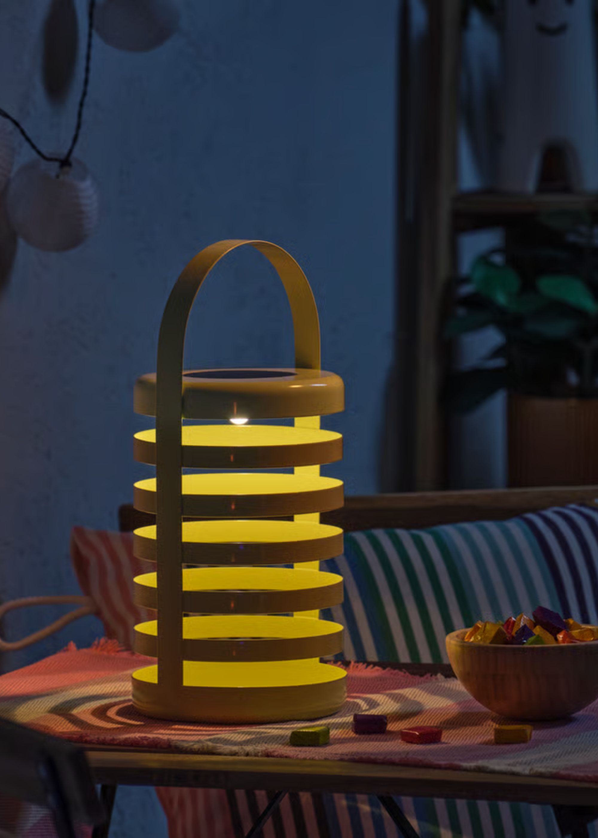 A light green open-striped design solar lantern with a handle turned on and sitting on top of an outdoor table at nighttime next to a bowl of sweets