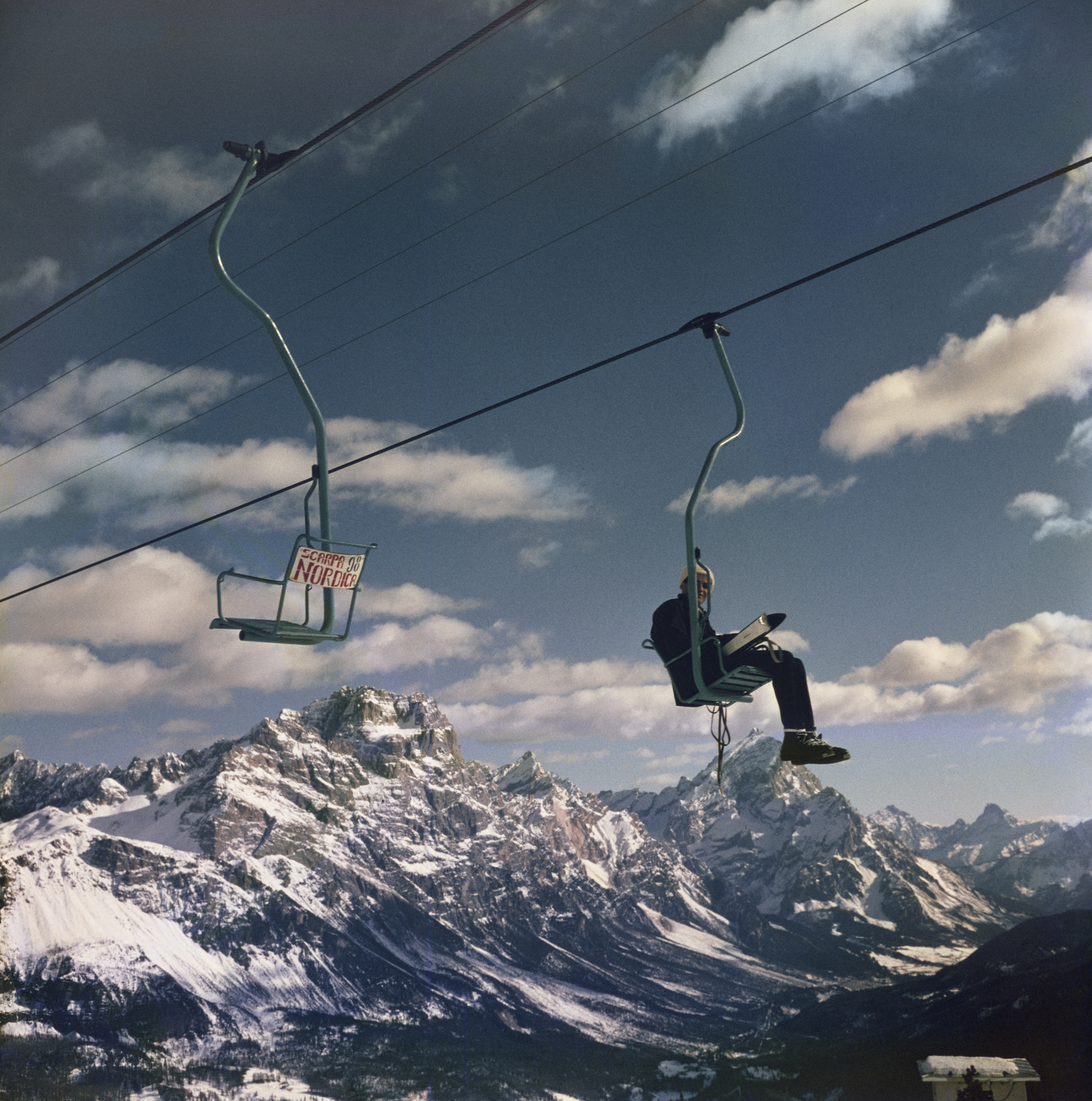 A lone skier riding an open chairlift high above a snow-covered alpine valley, with rugged mountain peaks stretching into the distance.