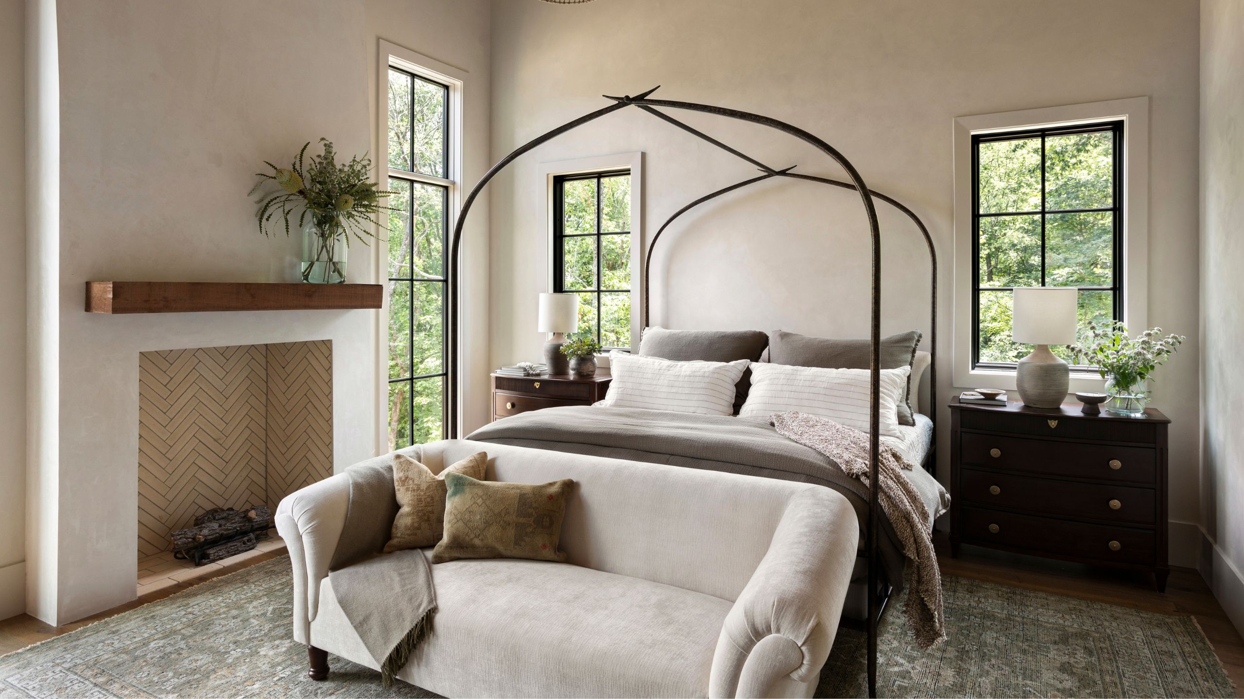 a neutral primary bedroom with large glass and black metal windows with a black metal four poster bed and soft neutral layers