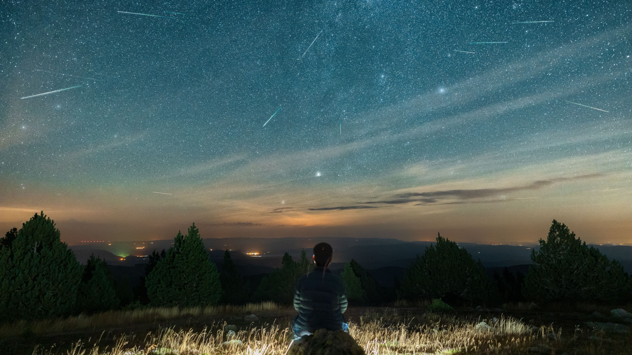 a person sits and looks at a sky full of stars, there are meteors streaking through the scene.