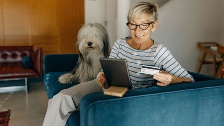 A chic, smiling woman shops online from her sofa with a credit card and tablet in her hands and a dog sitting beside her