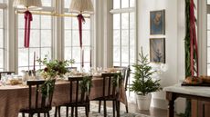 large bright neutral dining room with large windows decorated for christmas with a foliage garland, red ribbons hanging from the pendant lights, dark wooden chairs with garlands hung on the back, and a table laid for the holidays