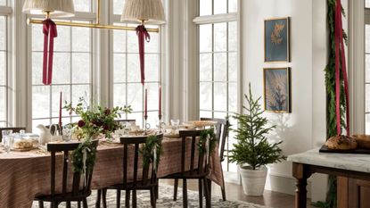 large bright neutral dining room with large windows decorated for christmas with a foliage garland, red ribbons hanging from the pendant lights, dark wooden chairs with garlands hung on the back, and a table laid for the holidays
