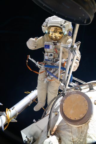 An astronaut in space, his golden visor reflecting a bent image of a space station module out of frame, gives a thumbs up. In the foreground immediately in front of him, a contraption of metal rods, similar to a folded camera tripod, extends from his mid section. Behind the astronaut, a thicker metal poll juts from out of frame, and below rests the corner of the air lock module.