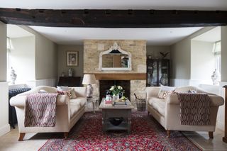 living room with matching cream sofas near exposed stone fireplace with woodburner and ceiling beams red traditional rug on pale stone floors
