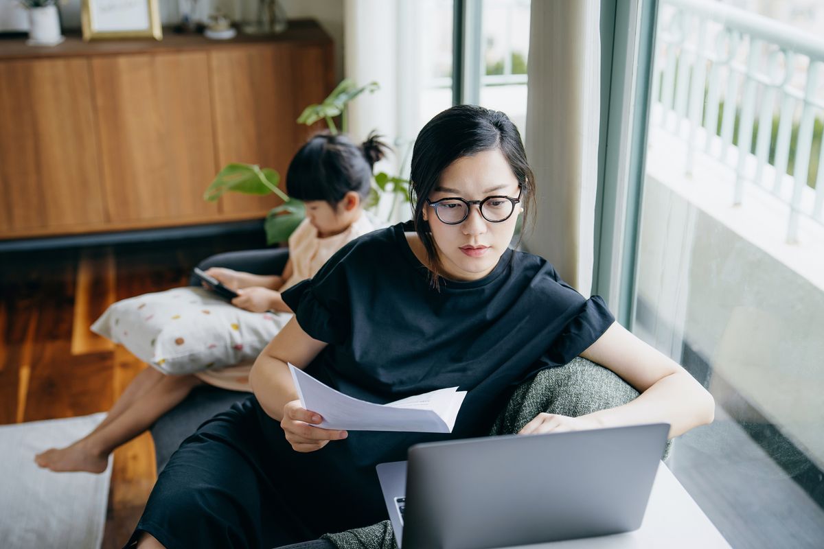 Asian mother working from home on laptop while little daughter is watching on digital tablet