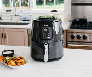 Ninja Air Fryer on a white kitchen counter with a plate of chicken nuggets and vegetables; in the background, a sink and stovetop.