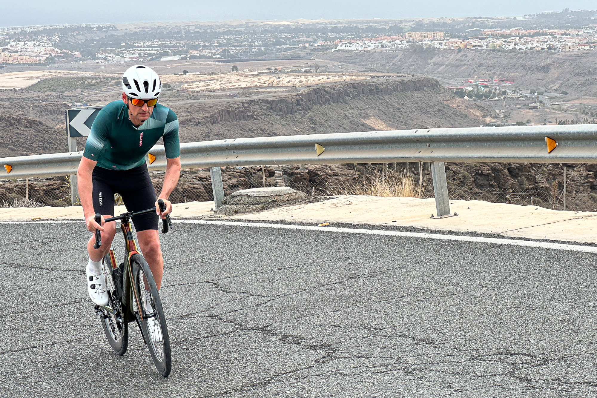 Man in a green jersey, black shorts and a white helmet riding an orange road bike up a hill out of the saddle in Gran Canaria