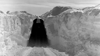 two walls of snow on either side show tire tracks on the ground leading to a dark tall tunnel.