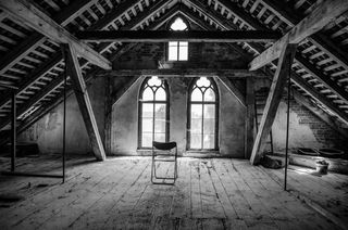 Black and white photo of wooden attic with cathedral-type windows and metal folding chair in center.